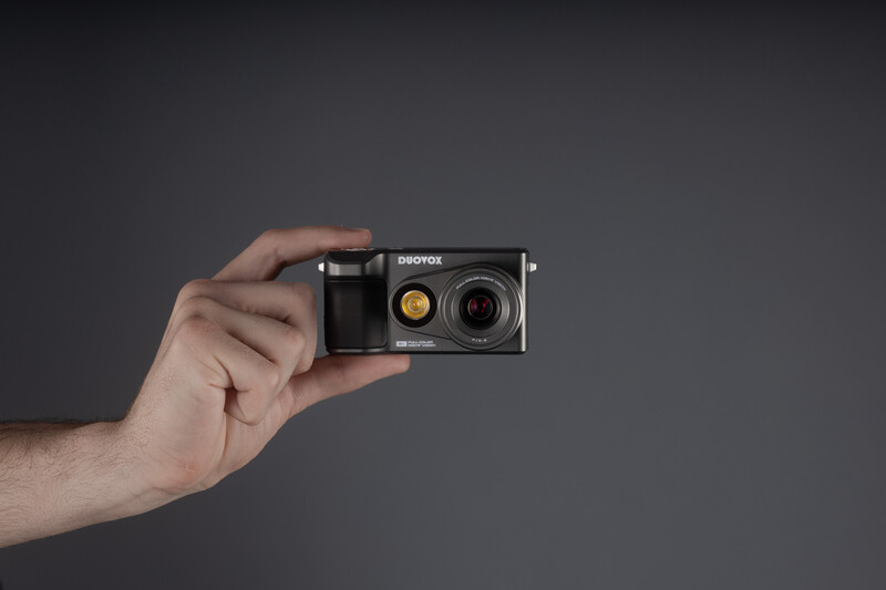 Hand holding a compact film camera with dual lenses against a neutral gray background.