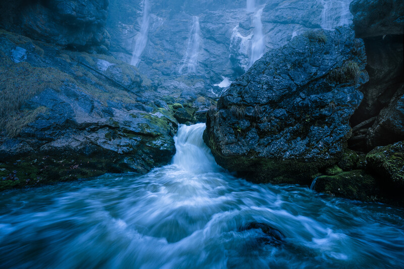 Rushing stream flows between moss-covered rocks in misty forest setting.
