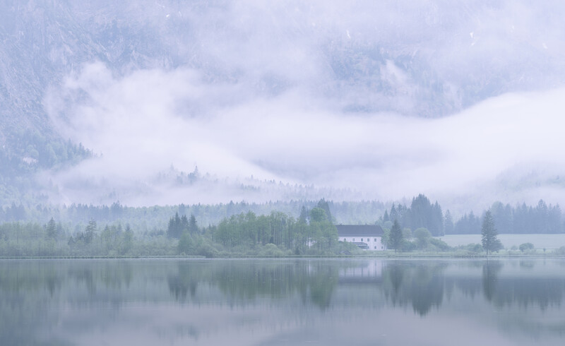 Misty lake landscape with forested shoreline and distant mountains under overcast sky.