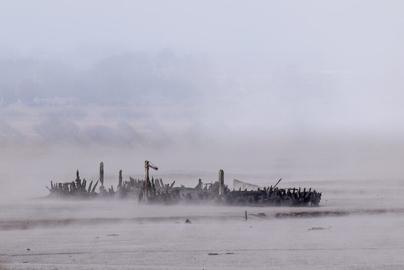 Silhouetted industrial structures emerging from dense fog over shallow water.