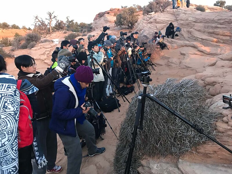 Large group of photographers with telephoto lenses gathered on rocky terrain during golden hour.