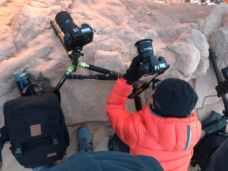Photographer in bright red jacket holding professional DSLR cameras above rocky terrain during golden hour.