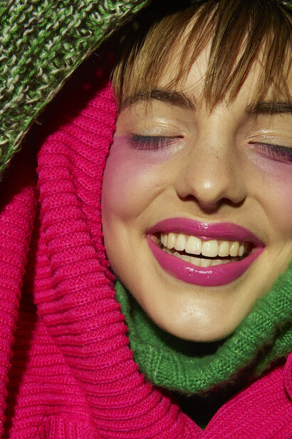 Close-up portrait of a smiling person with bright magenta lipstick and purple eyeshadow against a vibrant pink and green knit background.