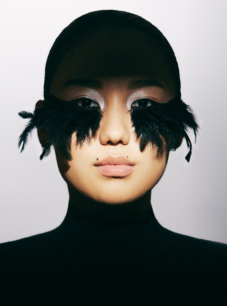 Portrait of person wearing black turtleneck and cap with dramatic black feathered eye covering against white background.