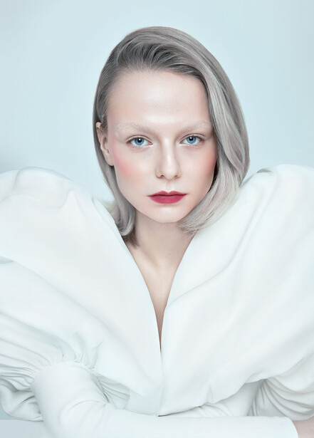 Portrait of a woman with platinum blonde hair wearing a white draped top against a light blue background.