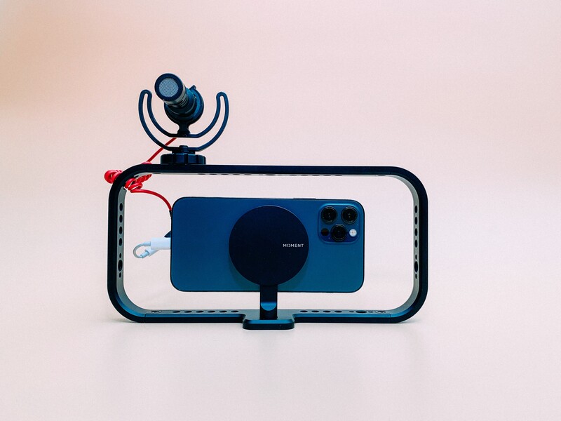 Smartphone mounted in a cage rig with microphone and lighting equipment against neutral background.