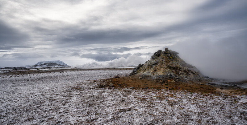 Rocky volcanic outcrop on barren, light-colored terrain under dramatic cloudy sky.