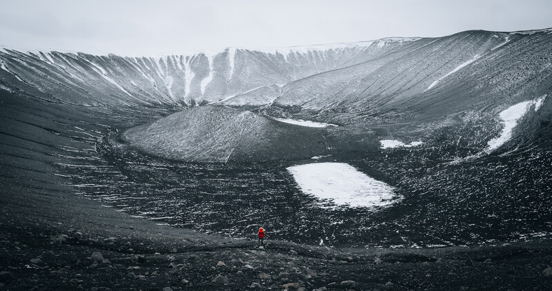Solitary figure in red jacket standing on dark volcanic terrain with snow patches and mountainous ridges in background.