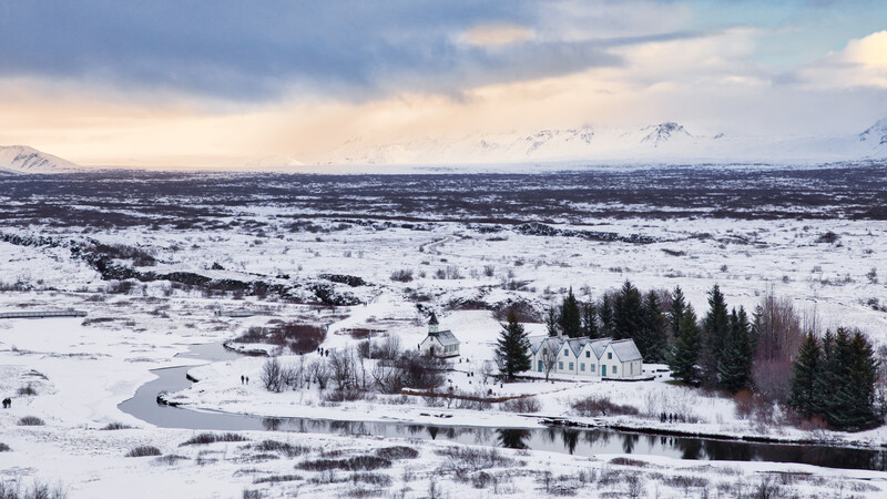 Snow-covered valley landscape with a white church beside a frozen river, evergreen forest, and snow-capped mountains in the distance.