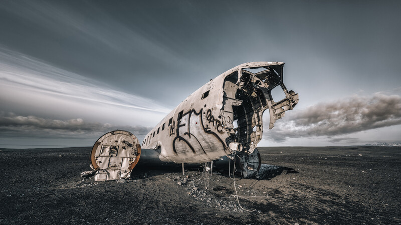 Wreckage of a large aircraft fuselage on a black sand beach under dramatic gray sky.
