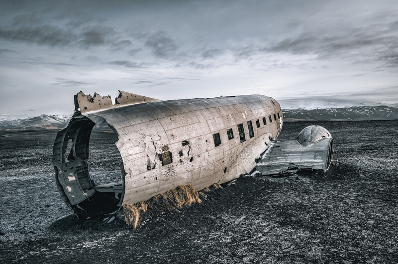 Wreckage of a large aircraft fuselage resting on a dark sand beach with mountains visible in the distance.