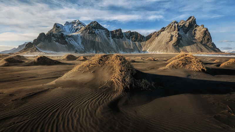 Black sand dunes with ripple patterns beneath jagged snow-capped peaks under blue sky.