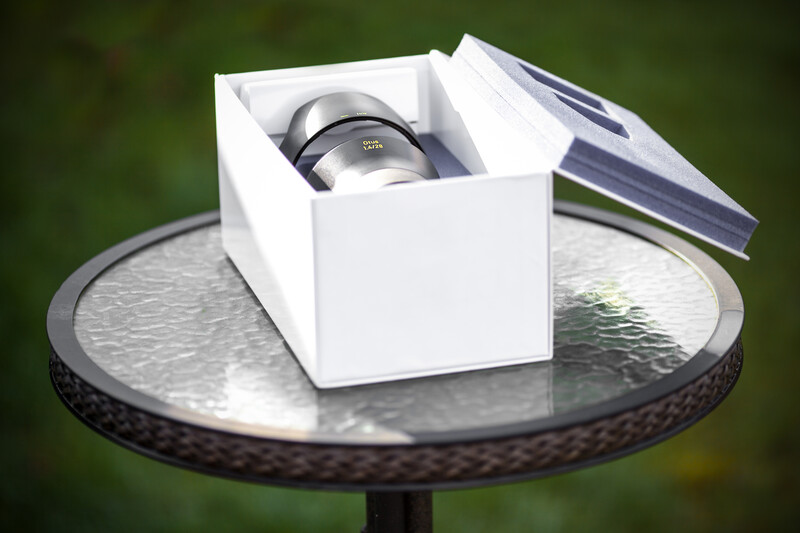 White product box with protective padding containing a telephoto lens, displayed on a reflective round table outdoors.