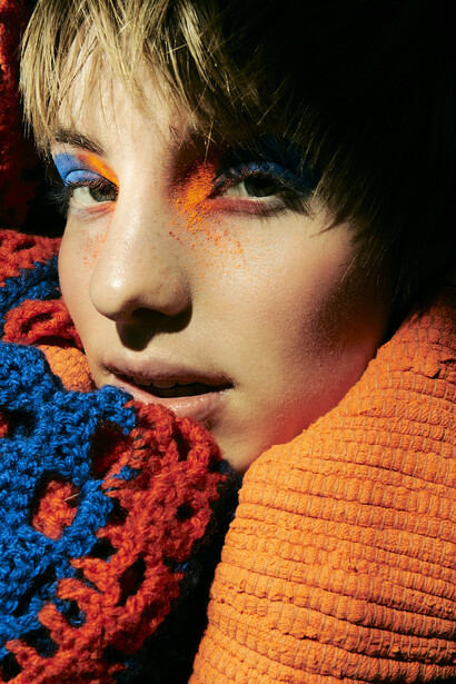 Child's face with vibrant blue and orange eyeshadow against colorful knit textures.