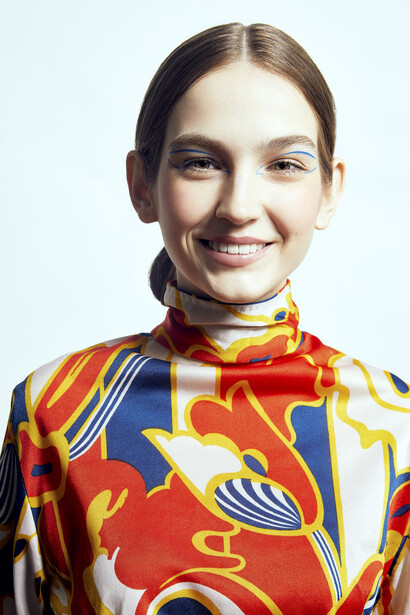 Young woman with straight hair smiling at camera wearing vibrant multicolored patterned turtleneck against plain light background.