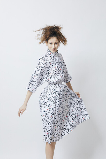 Woman in a patterned white dress with three-quarter sleeves turning to look over her shoulder against a plain white background.