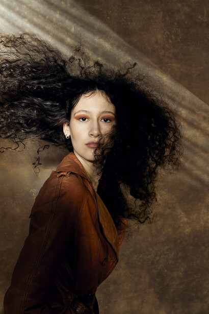Woman with voluminous curly dark hair wearing a rust-brown jacket, photographed against a textured neutral wall.
