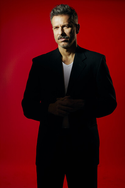 Man in black blazer and shirt against vibrant red background, hands clasped in front.
