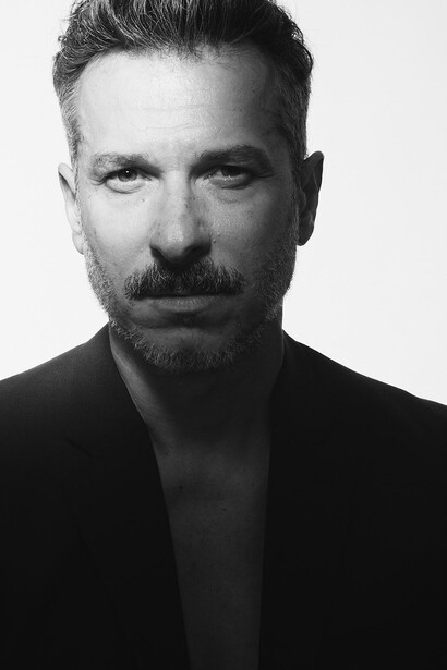 Black and white headshot portrait of a man with styled dark hair and facial hair, wearing an open dark shirt.