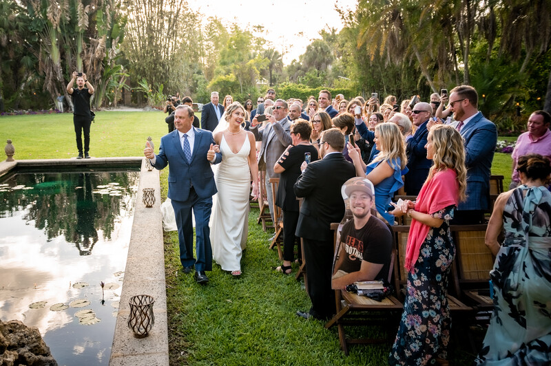 Bride and groom walking through gathered wedding guests on a manicured lawn beside a reflecting pond.