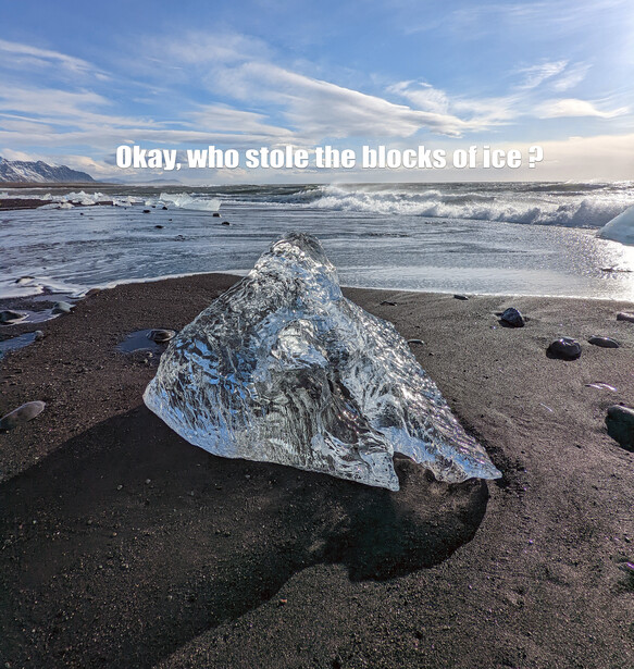 A translucent blue ice chunk rests on dark volcanic sand, with a frozen lagoon and mountains visible under a partly cloudy sky.