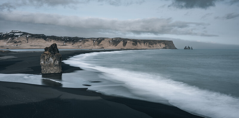 Solitary sea stack rises from black sand beach with white wave and distant cliffs under overcast sky.