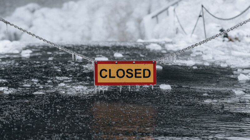 Red and yellow "CLOSED" sign attached to an icy wooden dock or pier structure, surrounded by frozen water and ice formations.