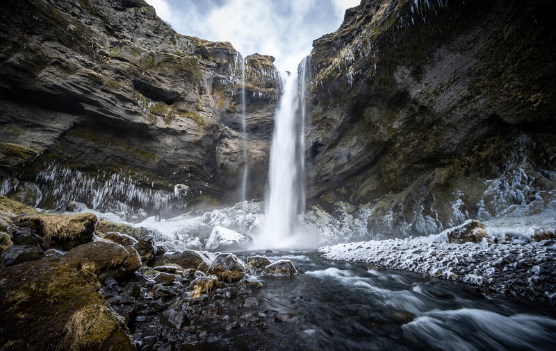 Frozen waterfall cascading between towering rock cliffs with icy formations and flowing water below.