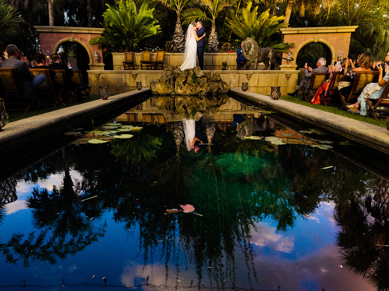 Bride and groom standing on a stone bridge over a reflecting pool at a formal garden venue during golden hour.