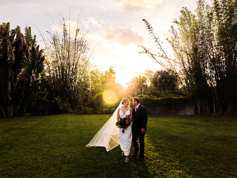 Bride and groom walking together on a grass lawn during golden hour, with tall ornamental grasses framing both sides.
