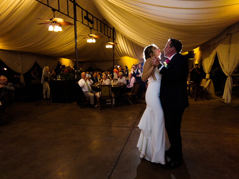 Bride and groom share their first dance under warm golden tent lighting with guests seated at tables in the background.