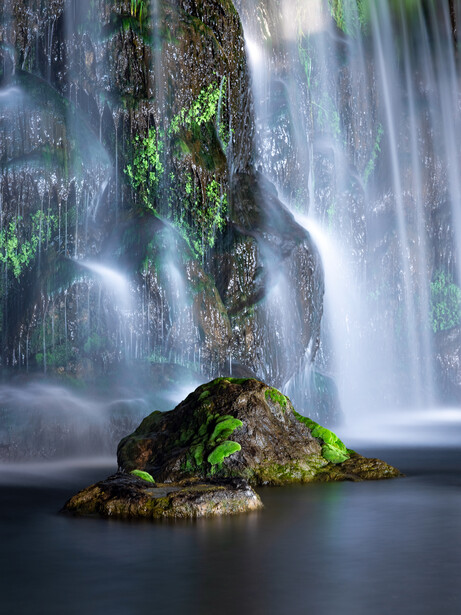 Moss-covered boulder in a stream surrounded by cascading waterfall with soft, flowing water.