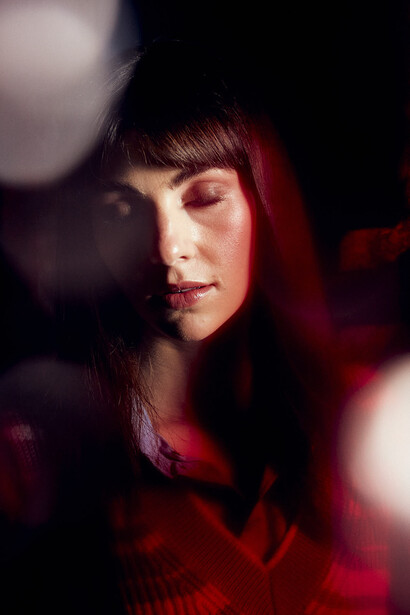 Woman with dark bangs and long hair lit by warm red and soft bokeh light against dark background.