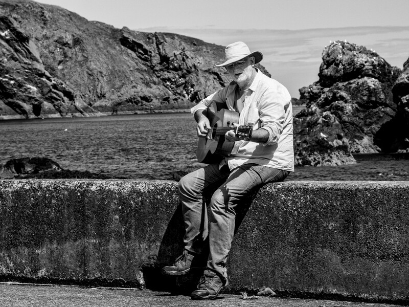 Man in white shirt and cowboy hat playing guitar while sitting on a concrete barrier overlooking a rocky coastline.