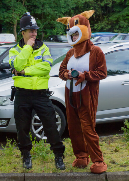 Police officer in high-visibility jacket standing next to person wearing brown animal costume in parking lot.