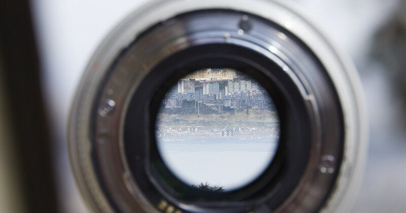 View through a telephoto lens showing a distant cityscape across water, with the lens barrel and focusing ring in sharp foreground focus.