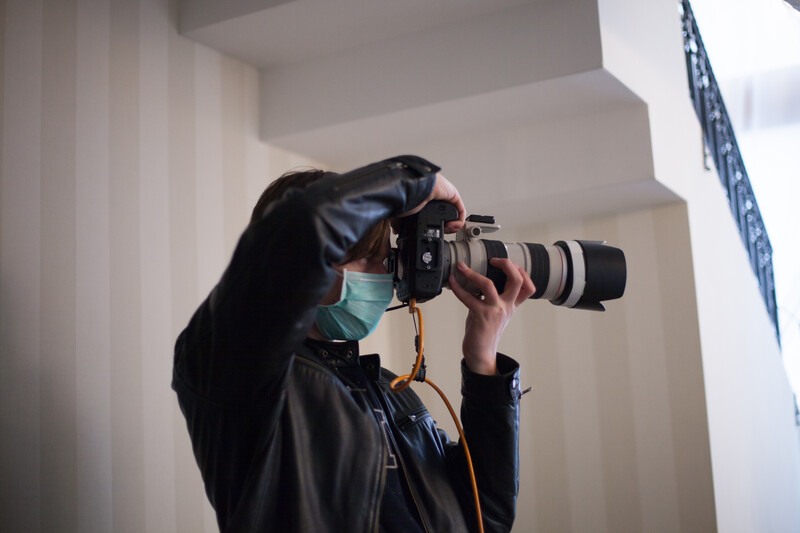 Photographer wearing a mask and dark jacket holding a DSLR camera with telephoto lens to their eye while shooting indoors.