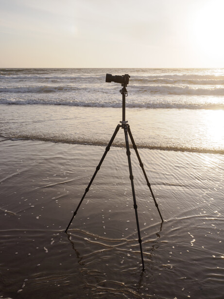 Camera mounted on tripod in shallow ocean water at sunset, casting long shadows on wet sand.