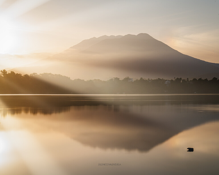Cone-shaped volcano reflected in still lake at sunrise, surrounded by mist and forested shoreline.