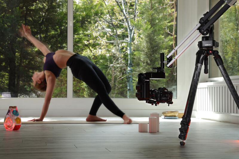 Fitness instructor demonstrating a downward dog yoga pose while being filmed with professional video equipment in a bright studio.