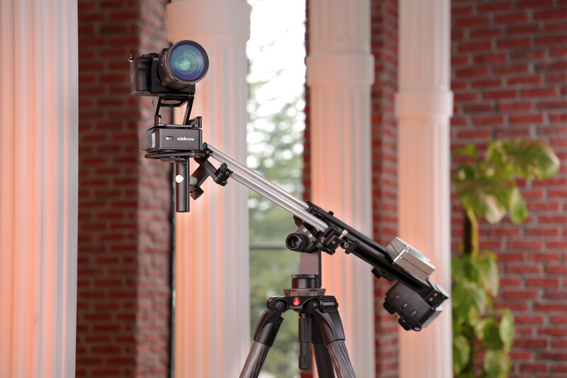 Camera mounted on jib arm with tripod over brick colonnade and green foliage.