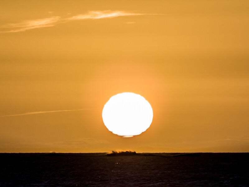 Bright sun sitting on the horizon at sunset over a dark ocean silhouette.