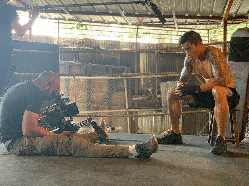Cinematographer operating a camera on shoulder rig while seated across from another person in an industrial space.