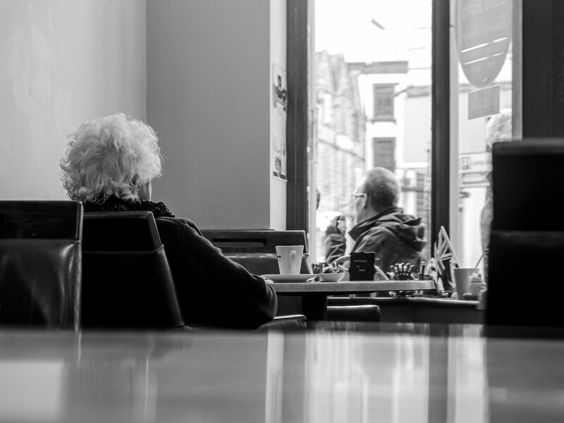 Elderly woman with white hair sitting at a table, viewing her reflection in a mirror or glass surface.