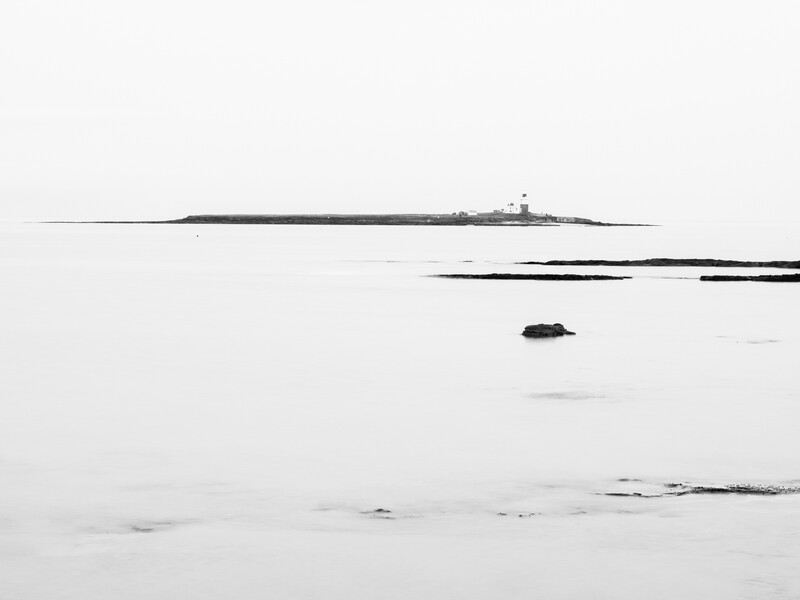 Minimalist seascape with a distant lighthouse and scattered rocks in calm, misty water.