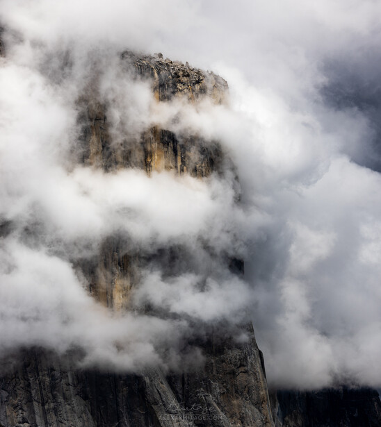 Dramatic mountain peak shrouded in thick clouds and mist, with rocky cliff faces barely visible through the fog.