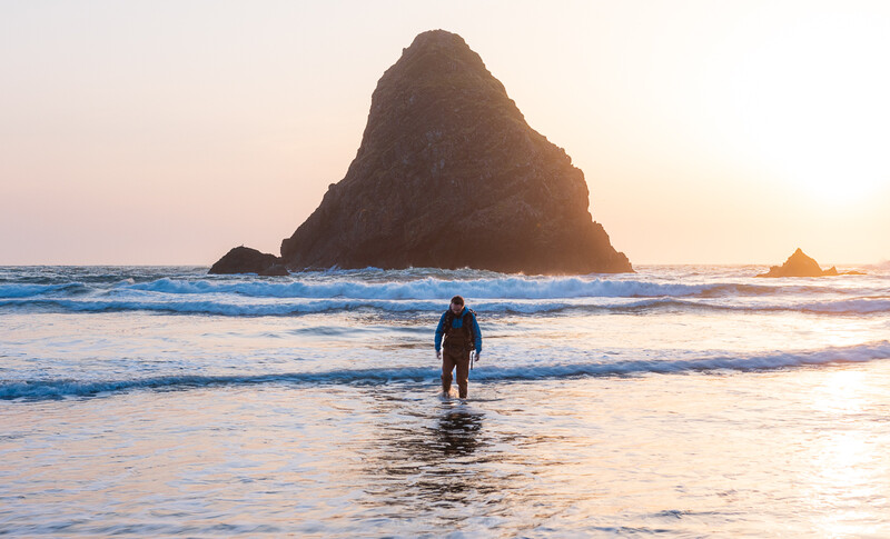 Surfer in wetsuit wading into ocean waves with towering sea stack in background at sunset.