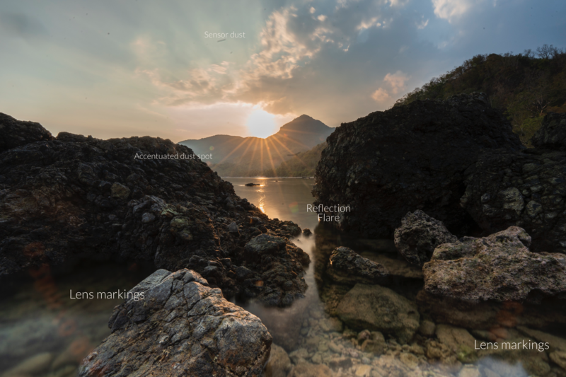 Rocky shoreline at sunset with sun rays breaking through clouds and mountain backdrop, annotated with photography terms.