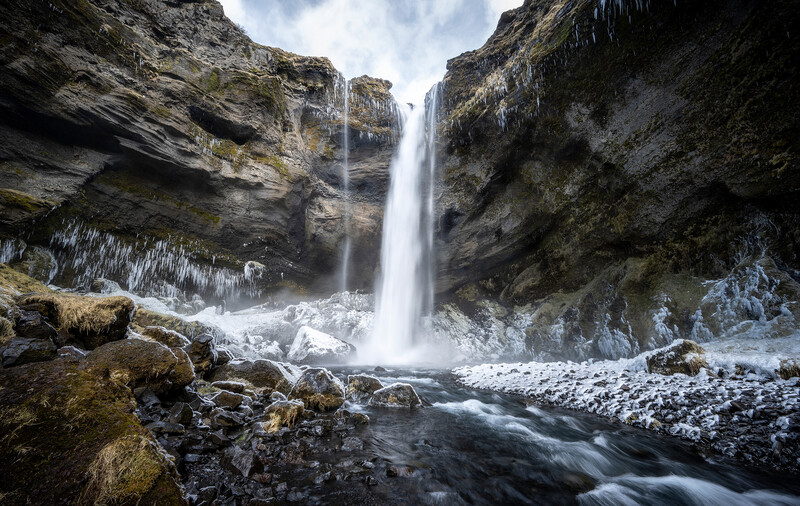 Waterfall cascading down moss-covered rock cliffs into a frozen stream below.