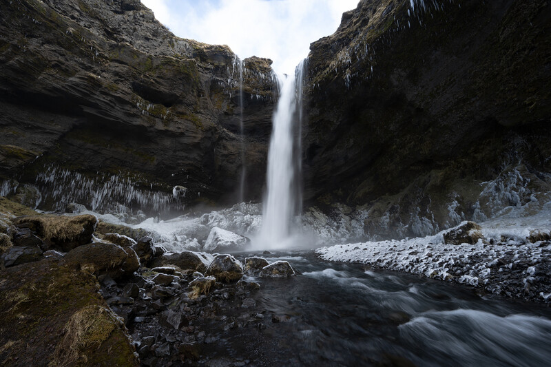 Waterfall cascading between dark rock cliffs into a misty pool with a flowing stream below.